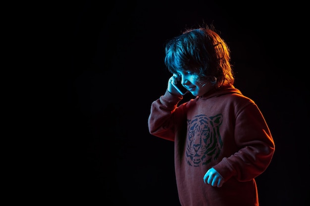 Lindo hablando por teléfono. Retrato de niño caucásico sobre fondo oscuro de estudio en luz de neón. Preciosa modelo rizada.