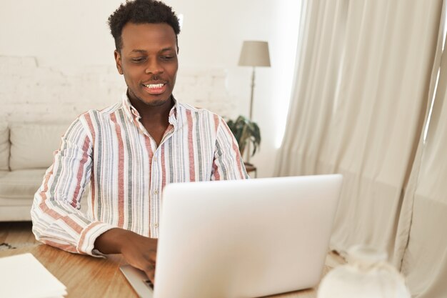 Lindo hombre africano joven relajado sentado usando una computadora portátil genérica para charlar con amigos, jugar videojuegos en línea, tener una mirada feliz