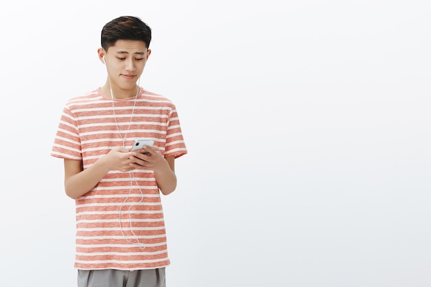 Lindo joven asiático en camiseta a rayas sosteniendo un teléfono inteligente con auriculares luciendo lindo y tocado en la pantalla del teléfono celular