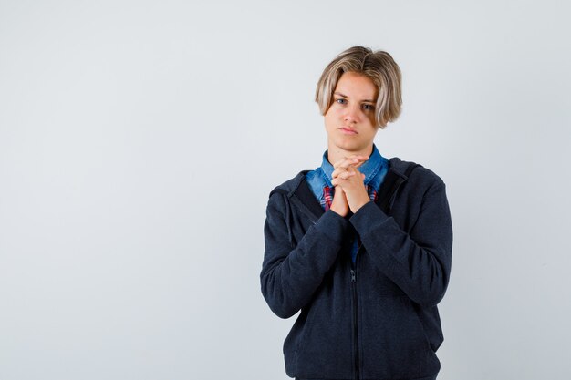 Lindo muchacho adolescente juntando las manos en gesto de oración en camisa, sudadera con capucha y mirando esperanzado, vista frontal.