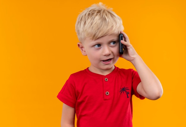Un lindo niño con cabello rubio con camiseta roja hablando por teléfono móvil en una pared amarilla