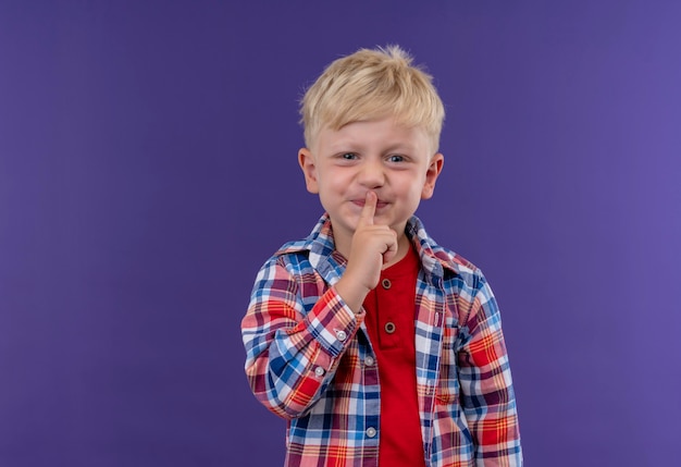 Un lindo niño con cabello rubio vistiendo camisa a cuadros manteniendo el dedo índice en la boca expresando shh gesto en una pared púrpura