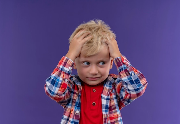 Un lindo niño con cabello rubio vistiendo camisa a cuadros manteniendo la mano en la cabeza sobre una pared púrpura