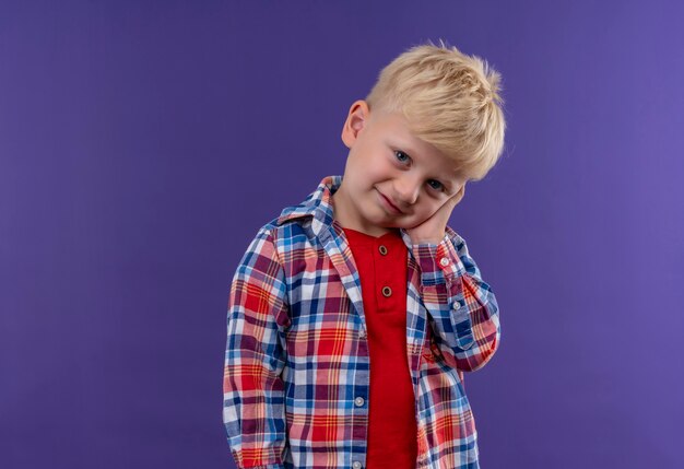 Un lindo niño con cabello rubio vistiendo camisa a cuadros manteniendo la mano en la cara mientras mira en una pared púrpura