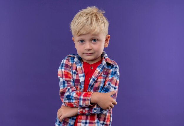 Un lindo niño con cabello rubio vistiendo camisa a cuadros mirando con las manos cruzadas sobre una pared púrpura