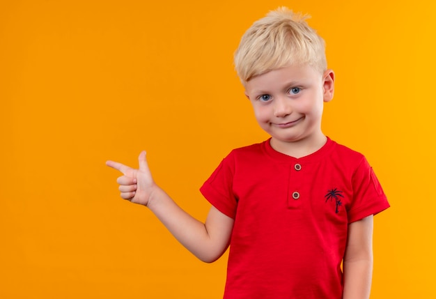 Un lindo niño con cabello rubio vistiendo camiseta roja apuntando a algo con el dedo índice mirando a una pared amarilla