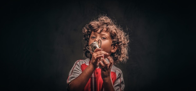 Lindo niño campeón en ropa deportiva con medalla de oro besando la copa del ganador. Aislado en un fondo de textura oscura.