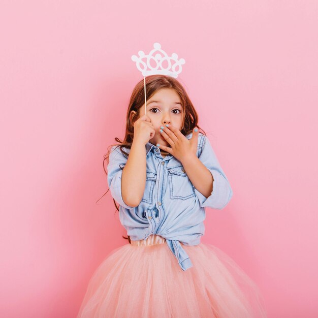 Lindo niño hermoso carnaval divirtiéndose aislado sobre fondo rosa. Niña bonita con cabello largo morena, falda de tul, con corona blanca en la cabeza expresando asombro a la cámara