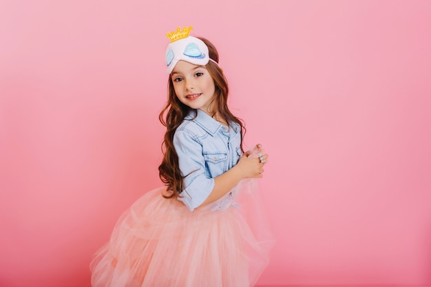 Lindo niño hermoso carnaval divirtiéndose aislado sobre fondo rosa. Niña bonita con cabello largo morena, falda de tul, máscara de princesa expresando felicidad a la cámara, celebrando la fiesta de los niños