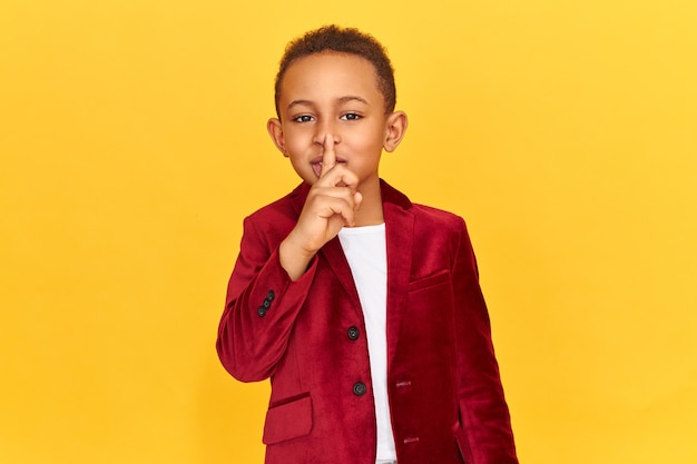 Foto gratuita lindo niño de piel oscura haciendo gesto de conspiración, manteniendo el dedo índice en los labios.