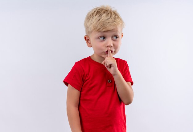 Un lindo niño rubio en camiseta roja que muestra el gesto shh con el dedo índice en la boca mientras mira de lado en una pared blanca