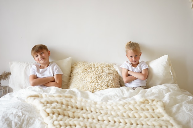 Lindo niño rubio relajándose en el dormitorio, sentado en la cama junto con su hermano mayor, ambos con los brazos cruzados y mirando a cámara y sonriendo. Niños, ropa de cama y concepto de hora de dormir.