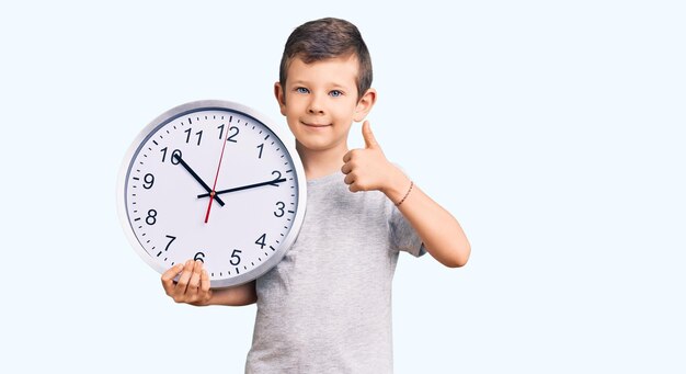 Lindo niño rubio sosteniendo un gran reloj sonriendo feliz y positivo pulgar hacia arriba haciendo excelente y señal de aprobación