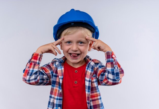 Un lindo niño sonriente con cabello rubio vistiendo camisa a cuadros en casco azul apuntando a su cabeza con los dedos índices mientras mira en una pared blanca