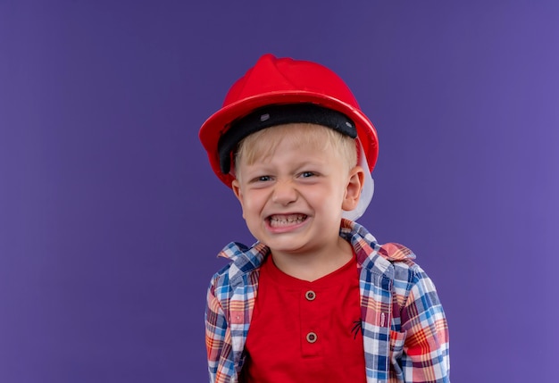 Un lindo niño sonriente con cabello rubio vistiendo camisa a cuadros en casco rojo mirando en una pared púrpura