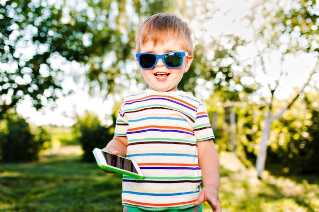 Lindo niño sosteniendo el teléfono en la mano y sonríe en el jardín de verano.