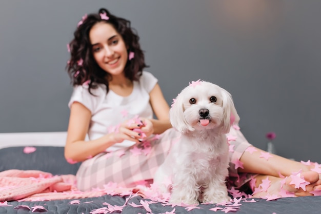 Lindo perrito encantador mostrando la lengua en la cama con una mujer joven y bonita. Relajarse en casa con animales domésticos, momentos divertidos.