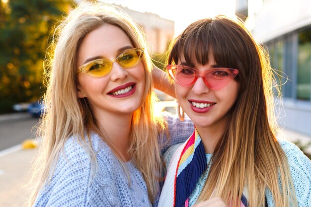 Lindo retrato soleado de cerca de dos magníficas damas muy elegantes sonriendo, vistiendo suéteres y gafas vintage, otoño primavera, metas de amistad.