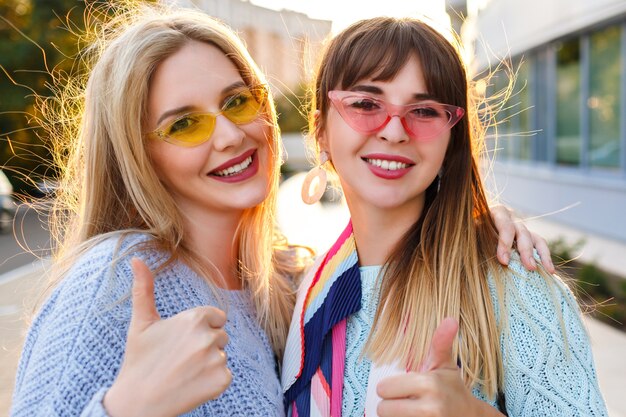 Lindo retrato soleado de cerca de dos magníficas damas muy elegantes sonriendo, vistiendo suéteres y gafas vintage, otoño primavera, metas de amistad.