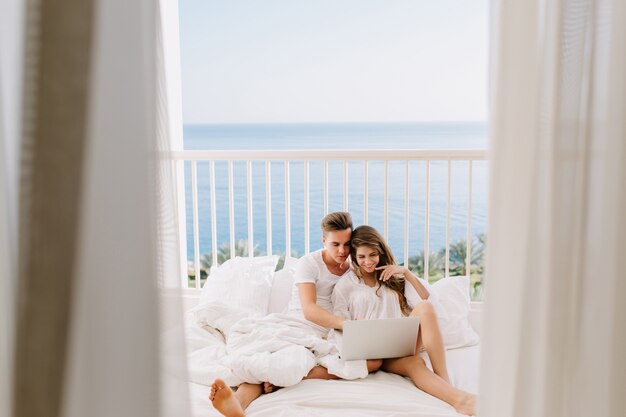 Lindos recién casados en ropa blanca sentados en la cama y viendo fotos de boda en la computadora portátil. Retrato de chico alegre descansando en la terraza con su hermosa novia con cortinas en primer plano