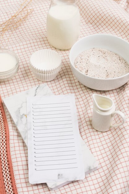 Lista en blanco en el diario con harina; Tarro de leche y moldes sobre fondo de tela.