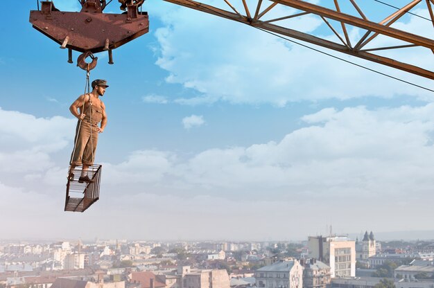 Listo para el día. Retrato de ángulo bajo de un joven constructor retro musculoso posando en un travesaño colgando de una grúa en un rascacielos