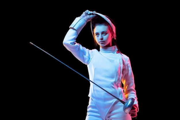 Listo. Jovencita en traje de esgrima con espada en mano aislada sobre fondo negro, luz de neón. Joven modelo practicando y entrenando en movimiento, acción. Copyspace. Deporte, juventud, estilo de vida saludable.