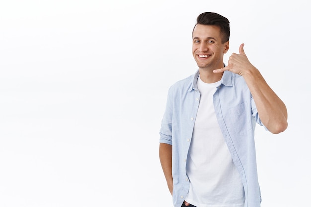 Llamame. Retrato de hombre adulto con estilo guapo que muestra la señal del teléfono cerca de la cara y sonriendo mientras promueve su servicio, dé el número de contacto en caso de que necesite hablar, de pie en la pared blanca