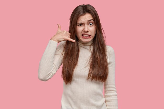 Llámame señal de vuelta. Mujer de cabello oscuro frustrada hace gesto de teléfono, levanta las cejas, viste ropa blanca