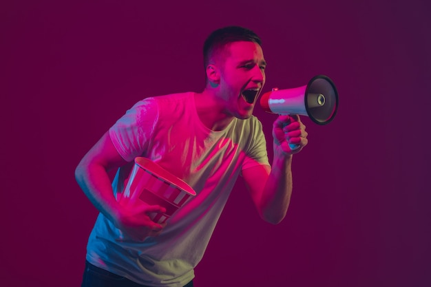 Llamar a ventas, gritar. Retrato de hombre caucásico aislado en la pared rosa-violeta en luz de neón. Modelo masculino con dispositivos. Concepto de emociones humanas, expresión facial,