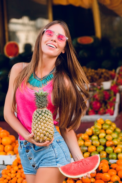 Lleno de alegría chica de verano divirtiéndose en el mercado de frutas tropicales. Ella sostiene ananas, rebanada de sandía y sonriendo