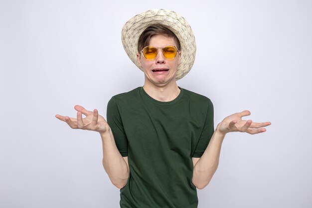 Llorando extendiendo las manos joven guapo con gafas con sombrero