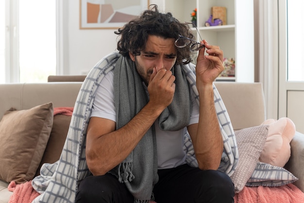 Foto gratuita llorando joven enfermo caucásico envuelto en tela escocesa con bufanda alrededor de su cuello sosteniendo lentes ópticos y poniendo la mano en la nariz sentado en el sofá en la sala de estar