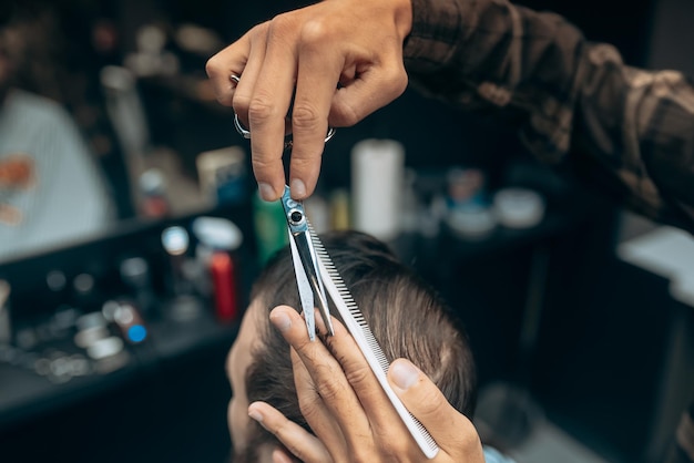 Lo pasamos genial en la barbería. Hombre barbudo joven alegre cortándose el pelo por peluquero mientras está sentado en una silla en la barbería