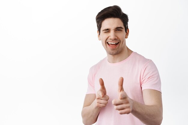 Lo tienes. Sonriente hombre guapo morena apuntando a la cámara con un guiño, animarle, apoyar a alguien, recoger, hacer un saludo amistoso descarado, de pie sobre fondo blanco