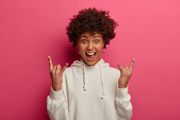 La loca chica de piel oscura se vuelve loca, asiste a un concierto de rock, hace letreros de heavy metal, exclama en voz alta, usa una sudadera, se siente entusiasmada y emocionada, usa una sudadera con capucha blanca, aislada en una pared rosa