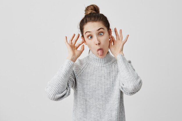 Loca joven divirtiéndose haciendo caras tontas. Diseñador de sexo femenino positivo entrecerrando los ojos posando con orejas que sobresalen. Concepto divertido