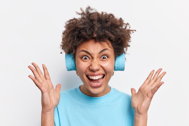 Loca mujer emocional mantiene las palmas levantadas grita en voz alta reacciona ante noticias increíbles vestida con una camiseta azul casual escucha música a través de auriculares aislados sobre fondo blanco Concepto de reacciones humanas