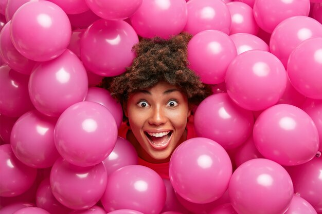 La loca mujer de piel oscura positiva se ve feliz, se sorprende, se divierte durante el día festivo rodeada de pequeños globos rosados inflados.