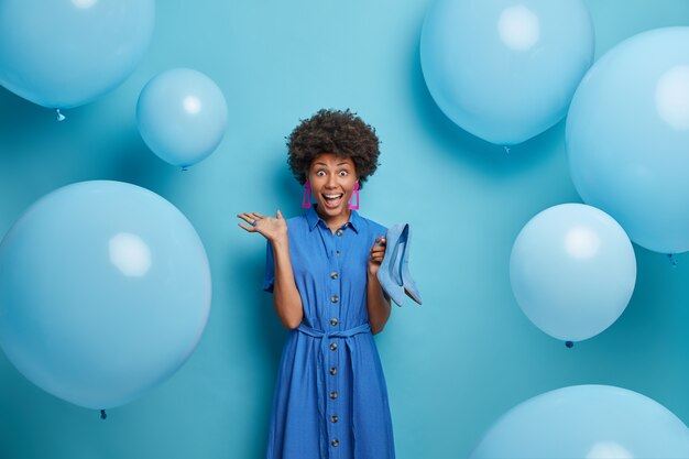 La loca mujer rizada emocional se ve feliz, feliz de recibir zapatos de tacón como regalo de su esposo, vestida con todo lo azul, globos inflados alrededor. Gente, vestimenta y concepto de fiesta.