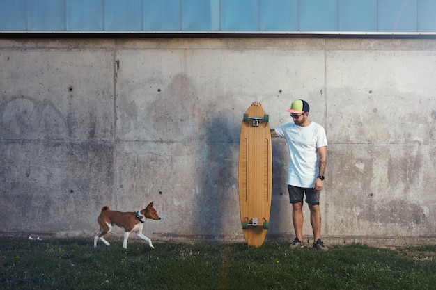 Longboarder barbudo y tatuado de pie junto a un muro de hormigón mirando un perro basenji marrón y blanco que se acerca