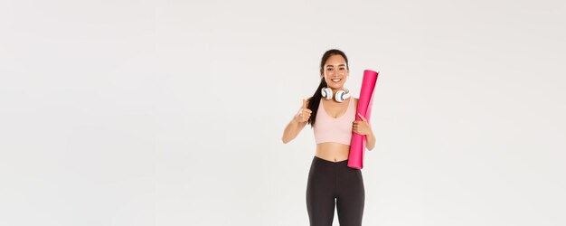 Longitud total de alegre chica asiática delgada satisfecha con alfombrilla de goma fitness y auriculares athl femenino