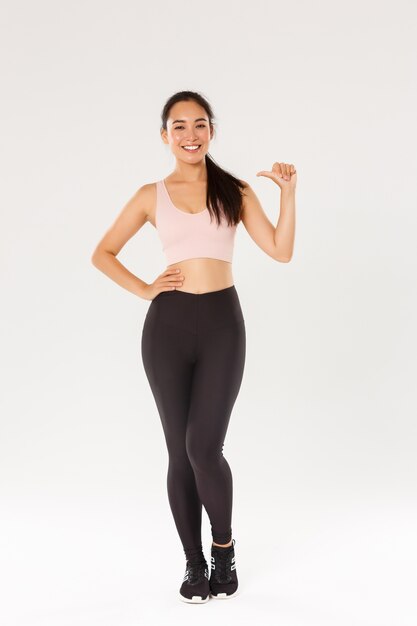 Longitud total de athelte femenino asiático sonriente confiado en sportsbra y leggigns, chica fitness apuntando a sí misma, llevando un estilo de vida activo y saludable, siendo profesional, de pie fondo blanco.