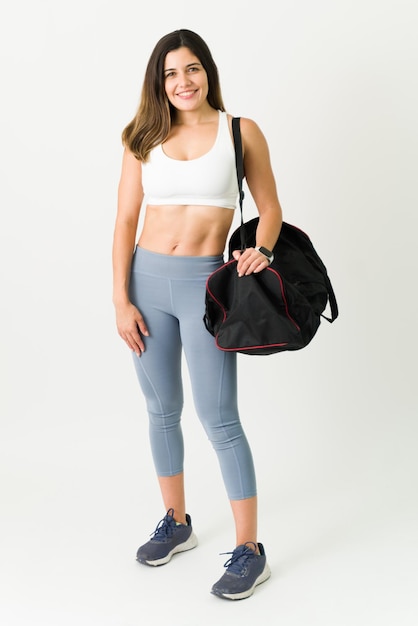 Longitud total de una atleta femenina atractiva que usa ropa deportiva sonriendo mientras lleva una bolsa de gimnasia. Seguro joven con abdominales tonificados