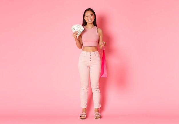 Longitud total de atractiva chica asiática sonriente en ropa de verano, sosteniendo dinero y una bolsa de compras, comprando algo caro, disfrutando de caminar en los centros comerciales, de pie en la pared rosa