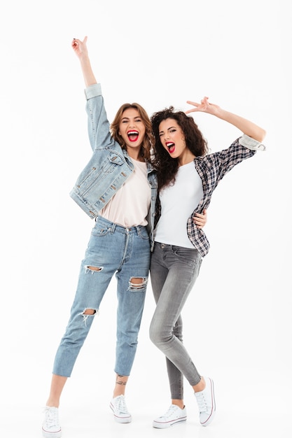 Longitud total dos alegres chicas de pie juntas y mostrando gestos de paz sobre la pared blanca