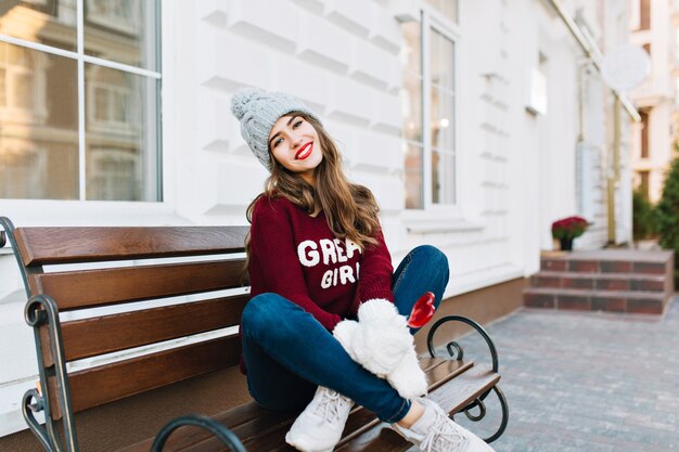 Longitud total hermosa joven con pelo largo en sombrero de punto, jeans y guantes blancos sentado en un banco en la calle. Ella tiene corazón de caramelo, sonriendo.