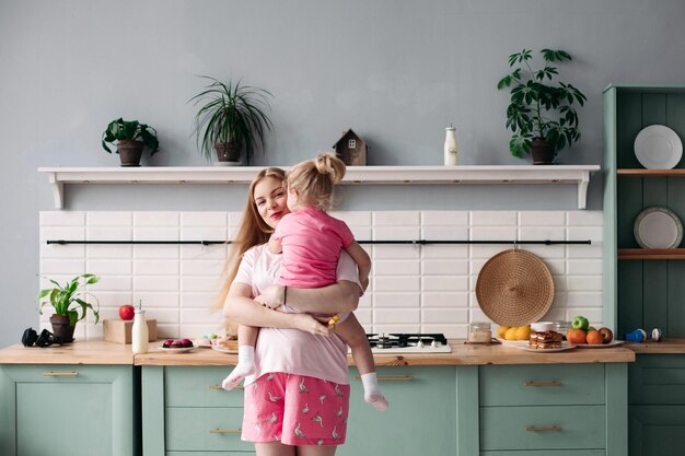 Longitud total de hermosa mujer sonriente en pijama sosteniendo a su encantadora hija en las manos y alimentándola con fruta para el desayuno Cocina moderna