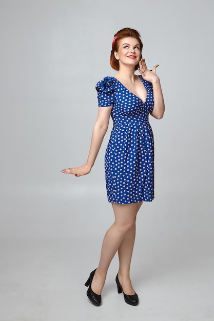 Longitud total hermosa pin up girl europea en elegante vestido azul con corte de cuello bajo con mirada tímida, sonriendo ampliamente mientras coquetea con alguien. Expresiones faciales humanas y cuerpo.