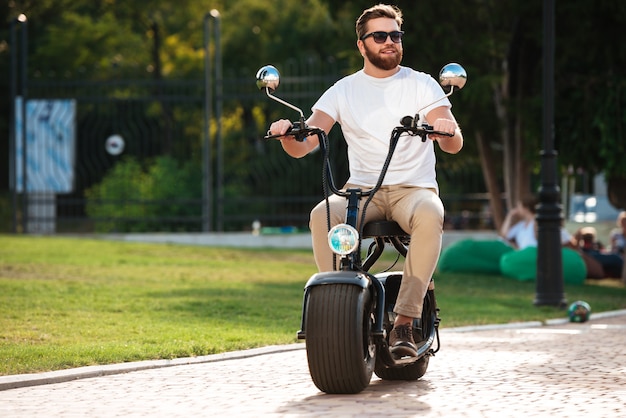 Longitud total del hombre barbudo feliz en gafas de sol paseos en moto moderna al aire libre y mirando a otro lado